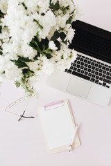 Office desk workspace with laptop, flowers bouquet and blank paper copy space mockup clipboard on pink table. Flat lat, top view.