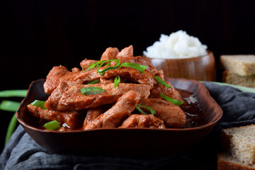 Pieces of meat stewed in tomato sauce with spices served in a clay bowl against black background