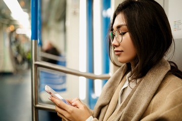 A beautiful business woman of Asian appearance in glasses sits in a subway car and looks at the screen of her smartphone