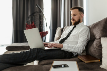 A bearded male freelancer in a shirt and tie is comfortably spread out on the couch, sitting in his living room, and working on a laptop