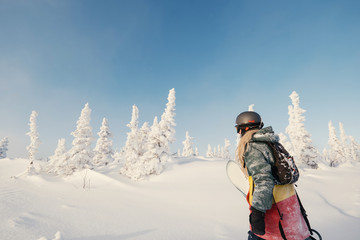 Female snowboarder wearing long dreadlocks and hoody holding a board, walking in snow-covered...