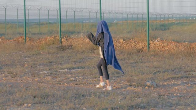 homeless child walks near protected area. skinny refugee girl head covered quilt goes along the iron fence state border. concept: war aftermath and political refugees.