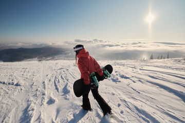 Woman snowboarder holding snowboard walking on mountain slop, preparing to snowboarding