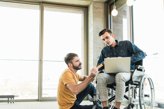 Young businessman in wheelchair and colleague using laptop in office