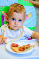little baby boy sits on a chair and a slice of pizza on a plate. unhealthy food