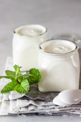 Two portions of natural homemade organic yogurt in glass jars with mint on a grey slate background. Fresh and natural fermented milk product.