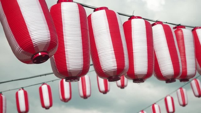 Many oriental paper red-white lanterns Chochin hanging on white cloudy blue sky background. Japanese lanterns hanging on bright sky background