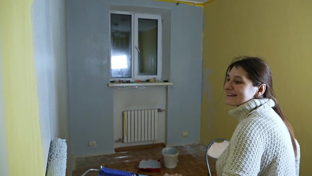 Joyful self-taught girl with a construction roller paints a yellow wall with gray paint. Independent apartment repair. Fresh repairs after moving to a new apartment