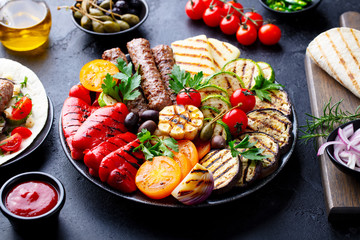Grilled meat kebabs, vegetables on a black plate with tortillas, flat bread. Slate stone background. Close up.