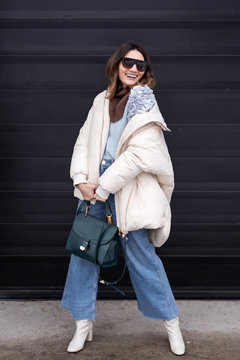 Caucasian girl in oversized down jacket, knitted sweater, flared jeans with a handbag and glasses posing on the street. Life style