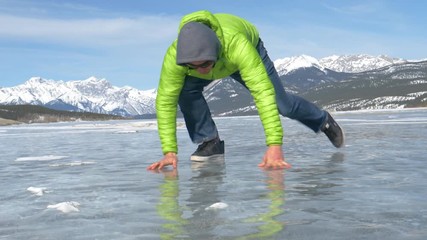 SLOW MOTION, LOW ANGLE, CLOSE UP: Funny shot of a young Caucasian man struggling to walk off the slippery surface of a frozen lake in the spectacular Canadian Rockies. Man slipping on icy Lake Abraham
