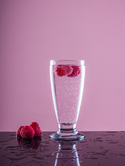 glass with sparkling water and raspberries on reflective surface with water and accompanied by raspberries