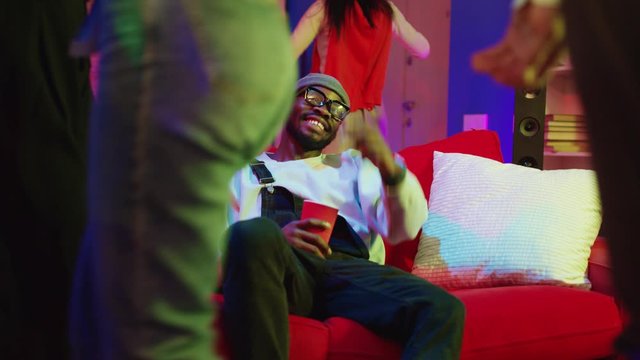 Funny drunk african american man drinking alcohol and relaxing on red couch flirting with girls dancing at home party. Nightlife. Happy people concept.