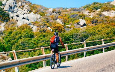 Bicycles on road at rocks of Capo Testa at Santa Teresa Gallura in Sardinia Island in Italy, summer. Biker driving on highway of Europe. Man Riding bike on motorway. Olbia province. Mixed media.