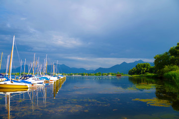 A small harbor on the Chiemsee lake (Bavaria, Germany) on a calm, beautiful and sunny summer day. Several sailboats are parked near a pier. Day is quite cloudy. Forests and mountains in the distance.