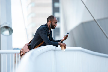 Smiling young businessman with earphones and coffee to go looking at smartphone