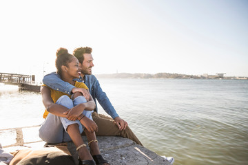 Young couple sitting on pier at the waterfront enjoying the view, Lisbon, Portugal