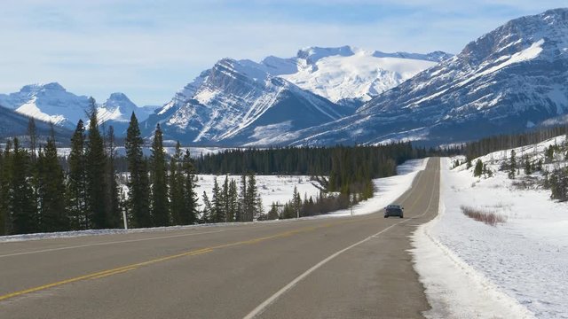 ICEFIELDS PARKWAY, ALBERTA, CANADA, MARCH 2019: Blue car drives down the empty road leading towards the snowy Canadian Rockies. Tourists on road trip cruise along the Icefields Parkway on a sunny day.