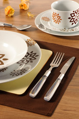 Table set up with dinnerware and silverware, ready for a meal. Close up of the fork and knife, with a set of dishes aside on a brown table mat, and cup and saucer on the background.