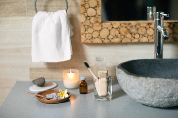 Natural bathroom accessories: bamboo toothbrush, make up remover in a glass container, coconut wax candle, organic bath salt, volcanic pumice and cotton towel standing near black stone sink.