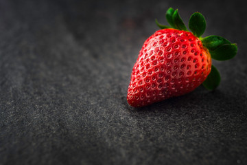 A close up macro photograph of a whole fresh ripe organic strawberry on a dark black slate stone background