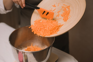 The cook adds the grated carrots to the metal bowl. Female hand put chopped carrot in wooden bowl with salad in kitchen. Cooking vegetables