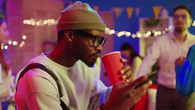 Handsome afro-american stylish man browsing app on smartphone drinking alcohol cocktail staying aside on birthday party celebration. Concept of nomophobia.