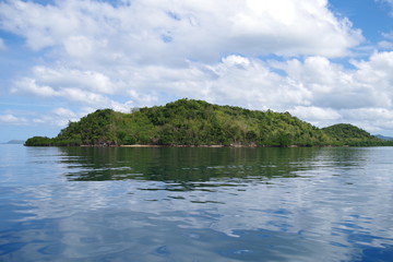 A small forested island in the Philippines archipelago. Isle is surrounded by the blue, calm waters of the ocean and azure, clear sky. There are few clouds, day is very bright and sunny.