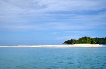 A shot of a very calm sea on a sunny day. There are few scattered clouds in the sky. On the horizon we can see a small island, overgrown with palm trees and sporting an inviting, sandy beach