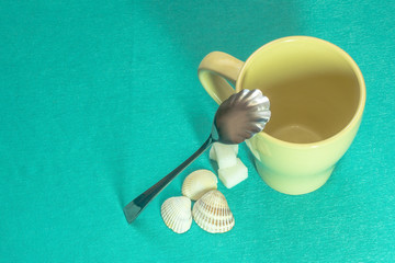 Tea set yellow cup, curly spoon, three pieces of sugar on a turquoise sea background decorated with shells