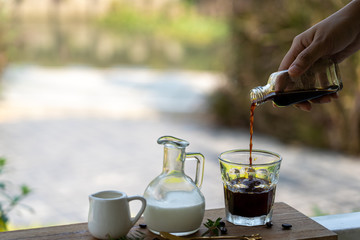 A woman's hand is pouring Espresso coffee in a clear cup. Morning coffee in a peaceful garden. With a milk jug and syrup on the side.