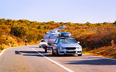 Car trailer with yacht or motorboat on road at Costa Smeralda in Sardinia Island, Italy summer. Minivan with motor boat on motorway on holidays at highway. Olbia province. Mixed media.
