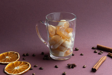 Glass cup with melting frozen coffee and milk on brown background with coffee beans, cinnamon sticks, dry orange slices