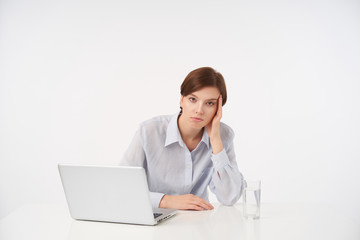 Exhausted young pretty short haired brunette lady with natural makeup leaning her head on raised hand and looking tiredly at camera, isolated over white background