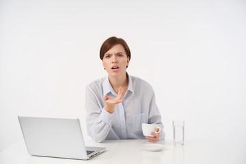 Excited young brown-eyed short haired brunette woman dressed in blue shirt frowning her eyebrows and raising confusedly palm while looking at camera, isolated over white background