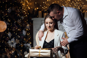 Husband gives his wife a gift. The wife sits on a chair. Wonderful young people in love. Woman holding Christmas present. Happy couple spending time together at home. Winter vacation. 