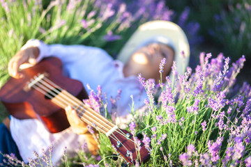 A country boy lies in a lavender field and plays the ukulele. The main subject is out of focus.