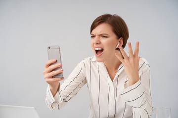 Emotional young pretty short haired brunette female with casual hairstyle frowning her face while shouting angrily over stressful video call, holding mobile phone while posing over white background