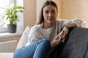 Fototapeta premium cute young girl in a white sweater and jeans sits on a sofa and looks at the camera