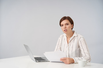 Bewildered young pretty short haired brunette lady with natural makeup sitting at table over white background and holding piece of paper, squinting while looking at camera with serious face