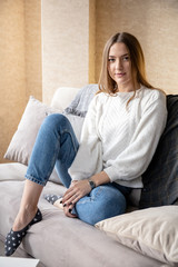 Obraz premium cheerful young girl in a white sweater and jeans with a lovely smile is resting on a sofa in a bright room