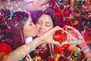 Fototapeta na wymiar Confetti party. Two young lesbian girls kiss and make a heart with their hands at a club party.