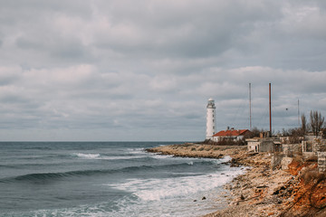 Beautiful seascape with a white lighthouse and old buildings on the shore. There is a wavy sea below, and a cloudy sky above