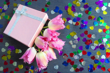pink fringed tulips stick out from a pink square-shaped cardboard box on a gray paper background multicolored foil hearts are scattered