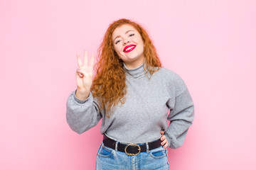 young red head woman smiling and looking friendly, showing number three or third with hand forward,...