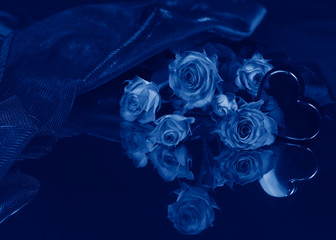 A bouquet of roses and an artificial heart lie on a mirror surface. Painted in the classic blue color trend 2020. Selective focus.