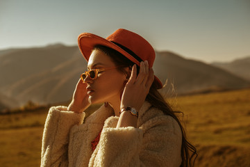 Outdoor close up fashion portrait of young beautiful confident brunette woman wearing stylish orange hat, sunglasses, wrist watch, faux fur coat, posing in mountain landscape. Copy, empty space  - obrazy, fototapety, plakaty