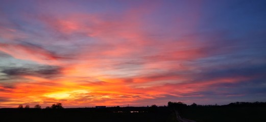 A fantastic sunset with red yellow orange and purple tones with dramatic cloud formation over green meadows with bare trees in January on the island of Rügen