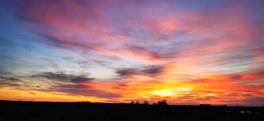 A fantastic sunset with red yellow orange and purple tones with dramatic cloud formation over green meadows with bare trees in January on the island of R&uuml;gen