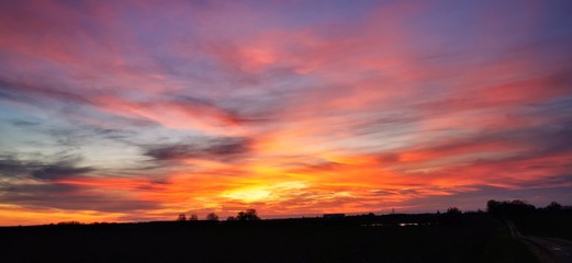 A fantastic sunset with red yellow orange and purple tones with dramatic cloud formation over green meadows with bare trees in January on the island of R&uuml;gen